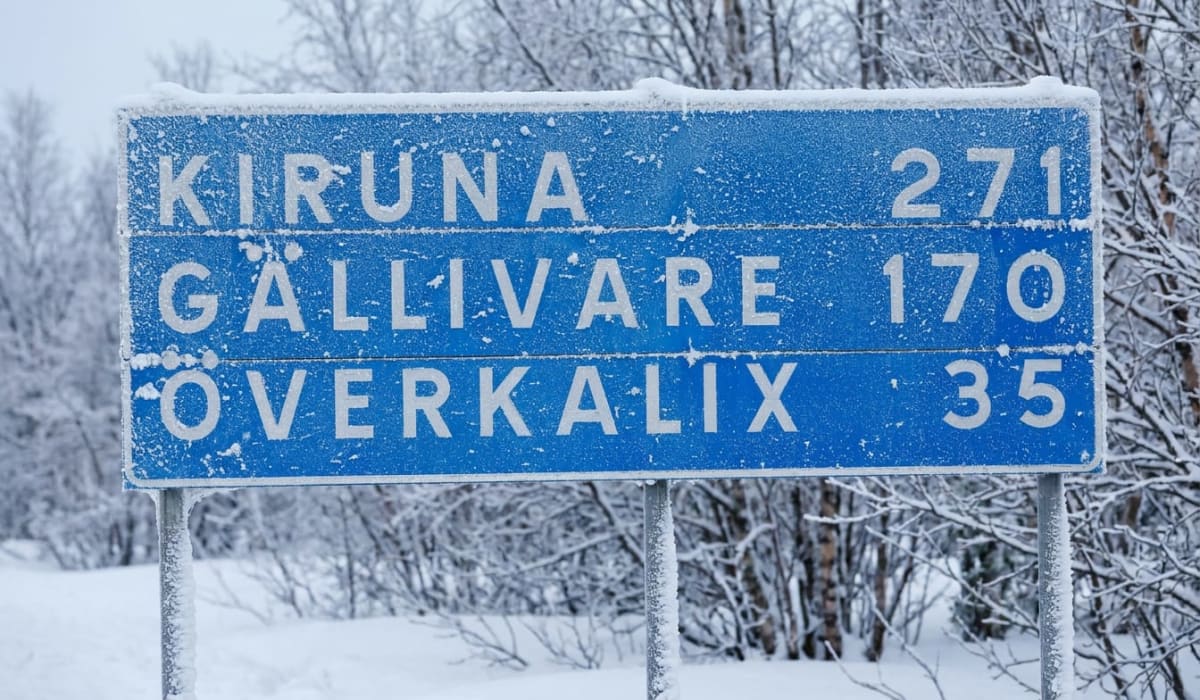En blå vägskylt med vit text som visar avstånd till Kiruna, Gällivare och Överkalix. Skylten och omgivande träd är täckta av snö och frost, vilket indikerar kallt väder.