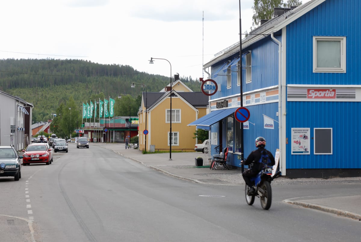 En gata med bilar parkerade vid sidan, en motorcykel kör bort från kameran, och byggnader med skyltar som "Sportia" och "Järn" längs vägen. I bakgrunden syns en skogsklädd kulle under en molnig himmel.