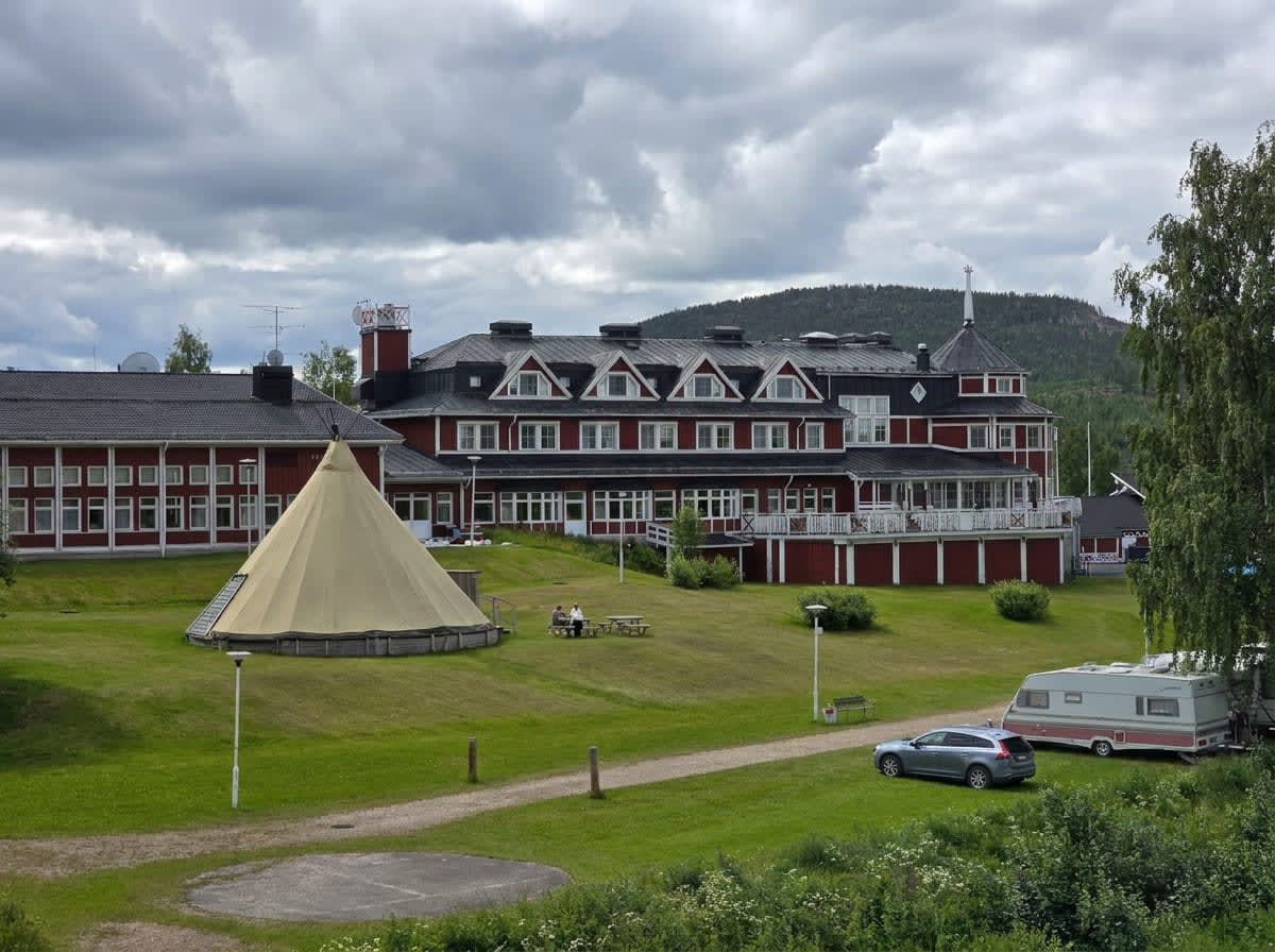 En stor, röd byggnad med flera våningar och vita knutar står mitt i bilden. Framför byggnaden finns en gräsklädd sluttning med ett beigefärgat tält, ett bord med två personer och en bil med husvagn. Himlen är mulen och i bakgrunden syns skogklädda be