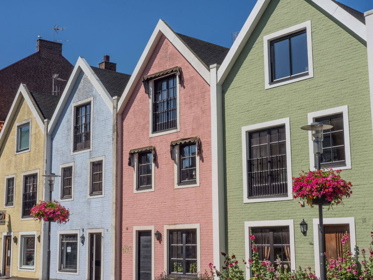 En bild som visar en rad färgglada husfasader i gult, ljusblått, rosa och ljusgrönt under klarblå himmel. Husen har sadeltak, vita fönsterramar och flera fönster med svarta spröjsar eller balkongräcken. En lyktstolpe med en blomsterarrangemang syns.