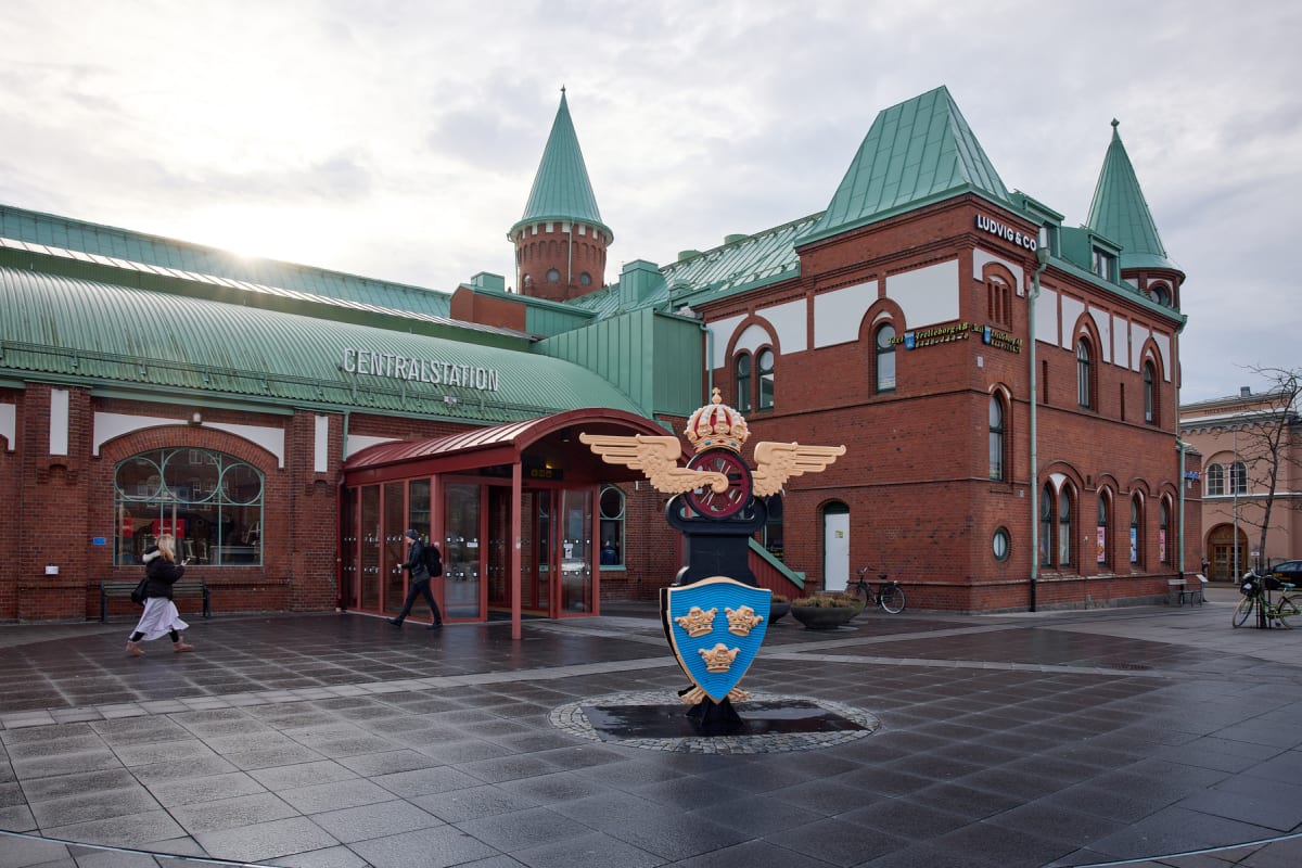En fasadvy av en röd tegelsbyggnad med ett grönt tak, där texten "CENTRALSTATION" syns. Framför byggnaden står en staty med vingar, en krona och tre kronor på en sköld. Två personer går på det våta stenbelagda torget.