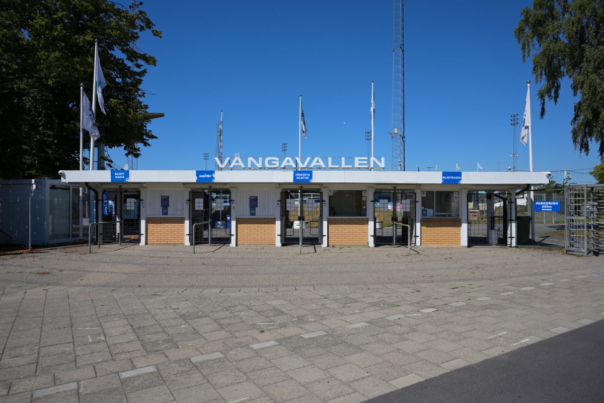En entrébyggnad till Vångavallen, med vita väggar och tegeldetaljer, syns under en klarblå himmel. Ovanför entrén står texten "VÅNGAVALLEN". Flera flaggstänger med vita flaggor och en radiomast syns i bakgrunden. Framför entrén ligger en grå stenlagd
