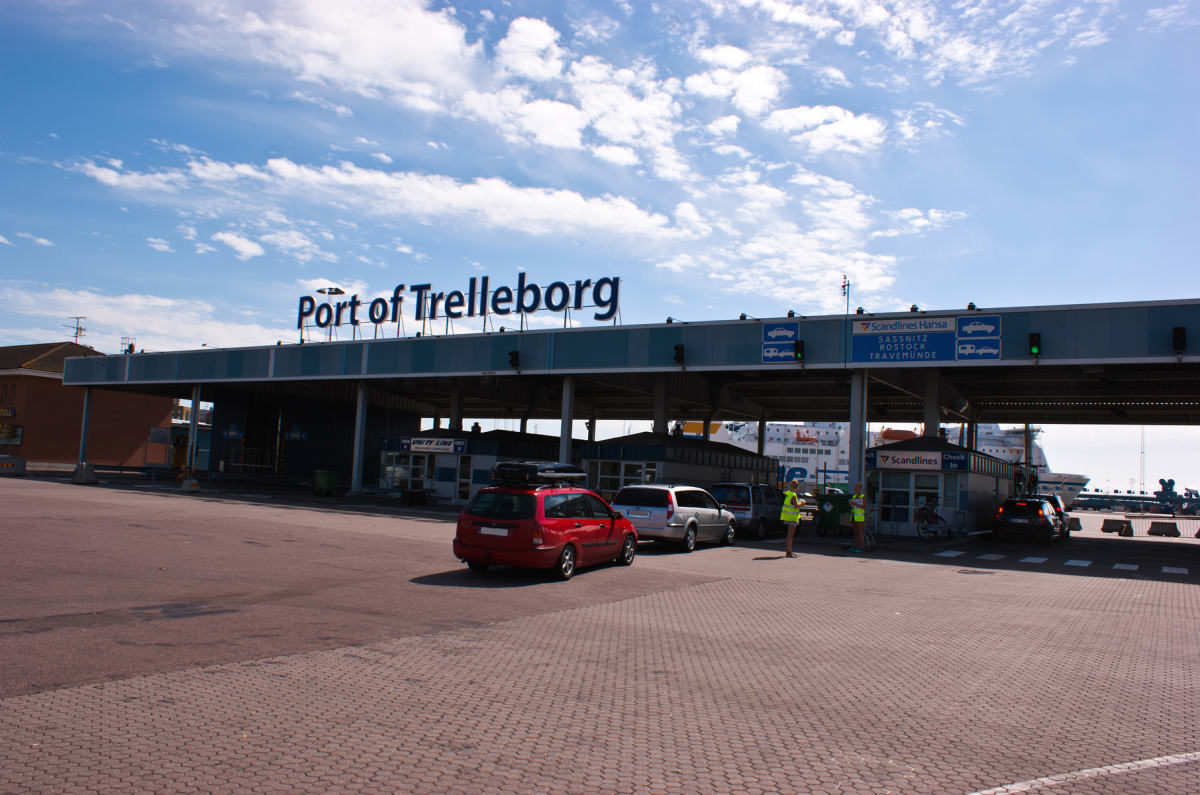 En bild av färjeterminalen i Trelleborg under dagtid. Flera bilar står i kö för incheckning och en stor färja syns i bakgrunden. En skylt med texten 'Port of Trelleborg' är placerad ovanför incheckningsbåsen. Två personer med gula västar står vid inc