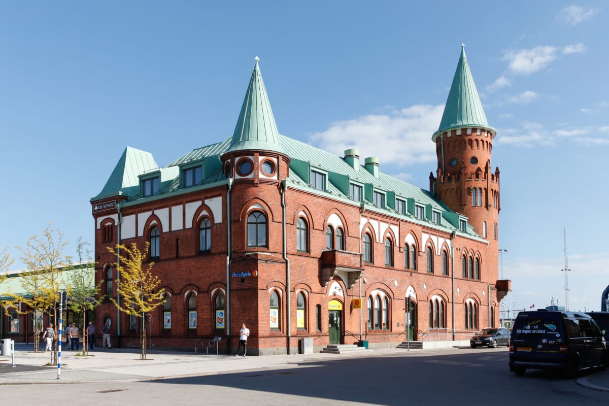 En stor röd tegelbyggnad med grönt tak och flera spetsiga torn står under en klarblå himmel. Framför byggnaden finns en asfalterad gata med parkerade fordon, och på trottoaren syns träd med vissna löv samt flera personer.
