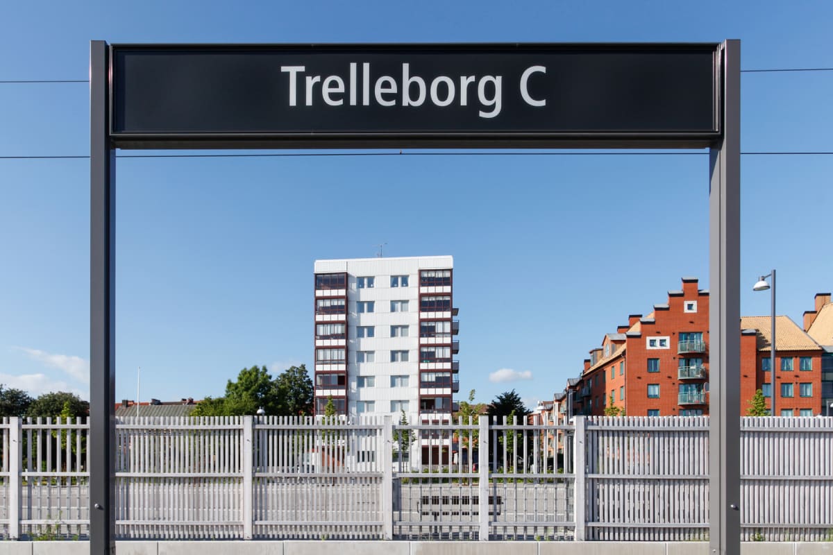 En svart skylt med vit text "Trelleborg C" sträcker sig över bildens övre del, stående på två grå stolpar. Bakom skylten och staketet syns en hög vit byggnad med balkonger och ett rött tegeltakshus, samt gröna träd under en klarblå himmel.