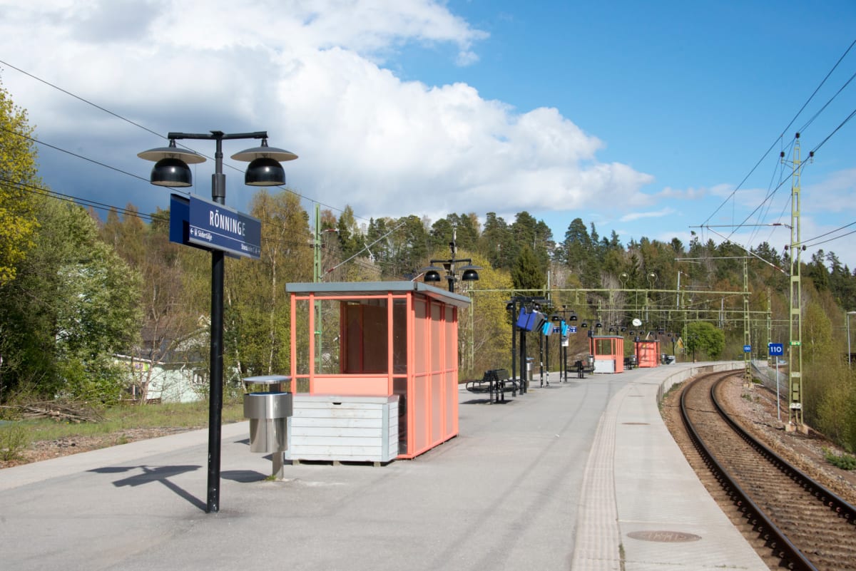 En bild av en tågperrong i Rönninge under dagtid med en spårkurva. På perrongen syns perrongtak, skyltar, soptunna, belysning och bänkar. Bakom perrongen finns en skogsklädd backe och delvis molnig himmel.