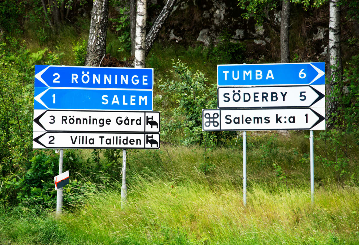 Två blå och vita vägskyltar står bland högt gräs med en bakgrund av grönska och träd. Skyltarna visar riktningar och avstånd till orter som Rönninge, Salem, Tumba och Söderby.