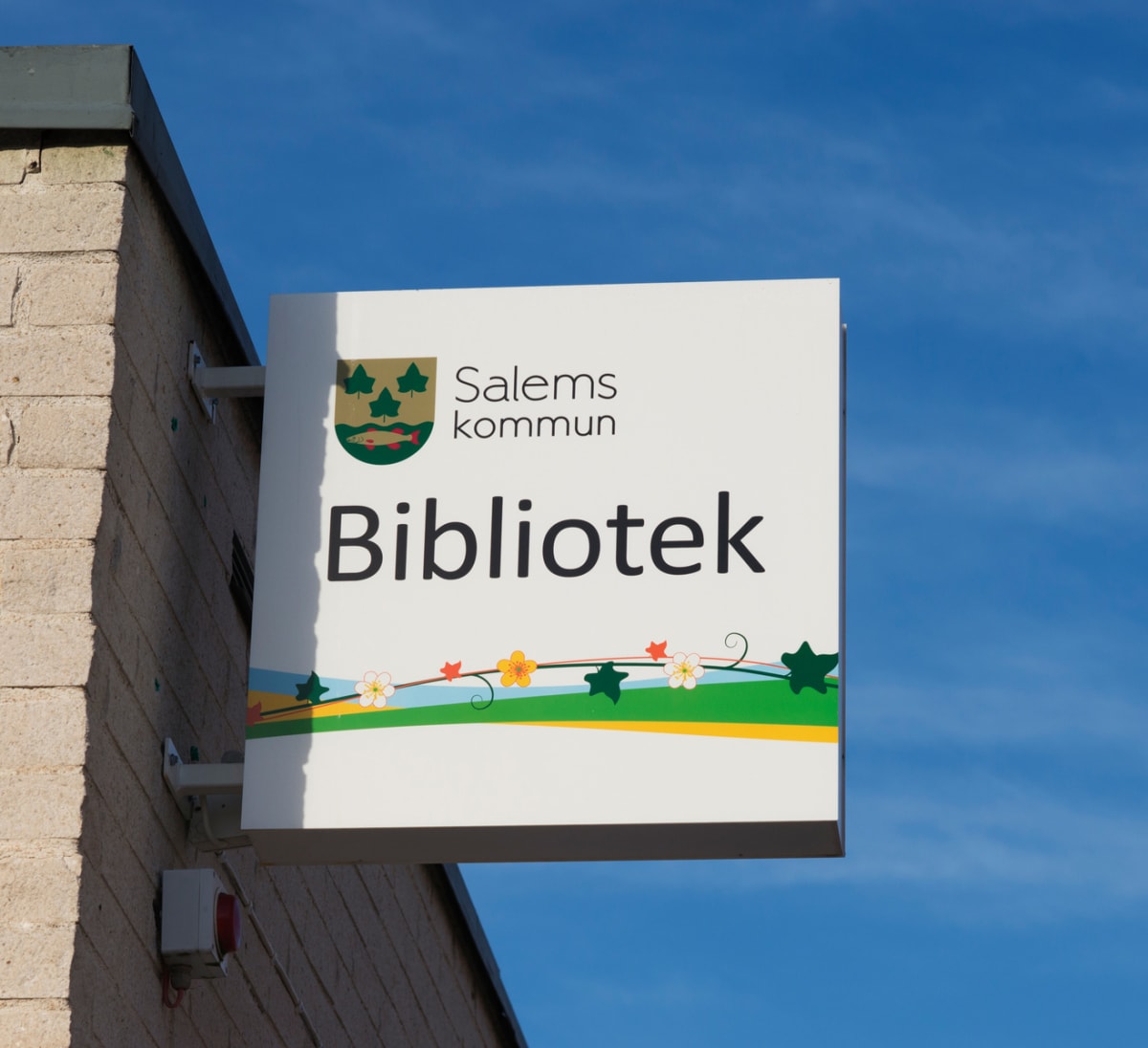 En fyrkantig skylt med texten "Salems kommun Bibliotek" hänger på en tegelbyggnad. Logotypen för Salems kommun, som består av gröna löv och en fisk på guldbakgrund, syns också på skylten. Under texten finns en dekorativ remsa med blommor och blad. Ba