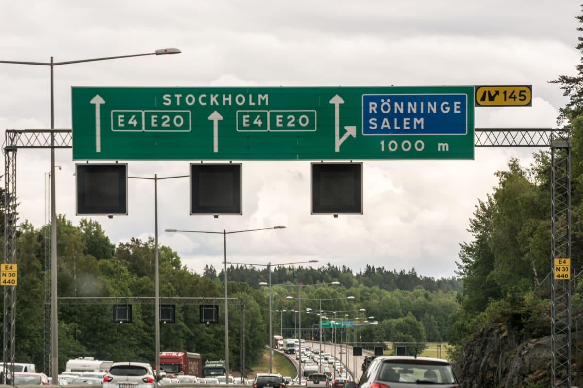 En stor grön vägskylt med information om riktning mot Stockholm (E4 E20) och avfart mot Rönninge/Salem (1000 m, väg 145) hänger över en motorväg med tung trafik. Tre svarta, rektangulära bildskärmar är monterade under skylten. Molnigt väder och grön