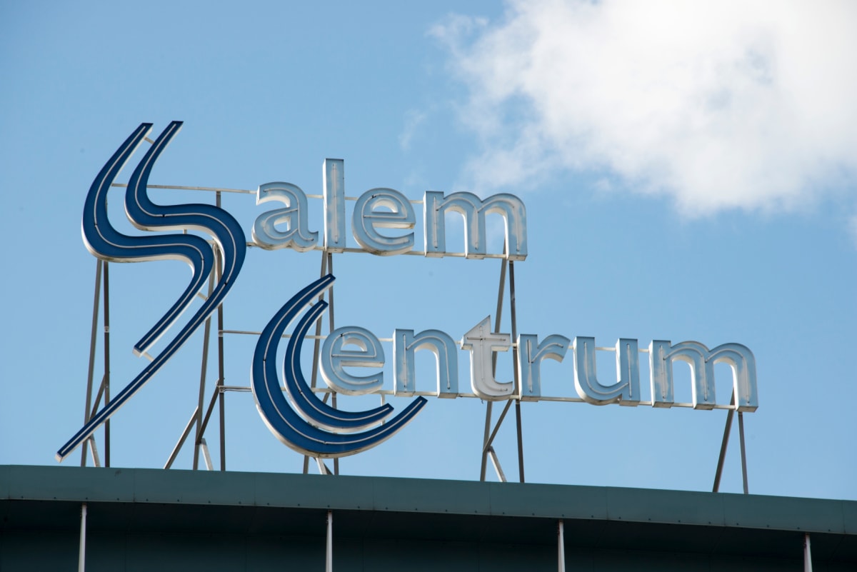 En bild som visar en del av en byggnad med en stor skylt med texten "Salem Centrum" i blå och silverfärgade bokstäver mot en blå himmel med vita moln. Skylten är monterad på en metallstruktur.