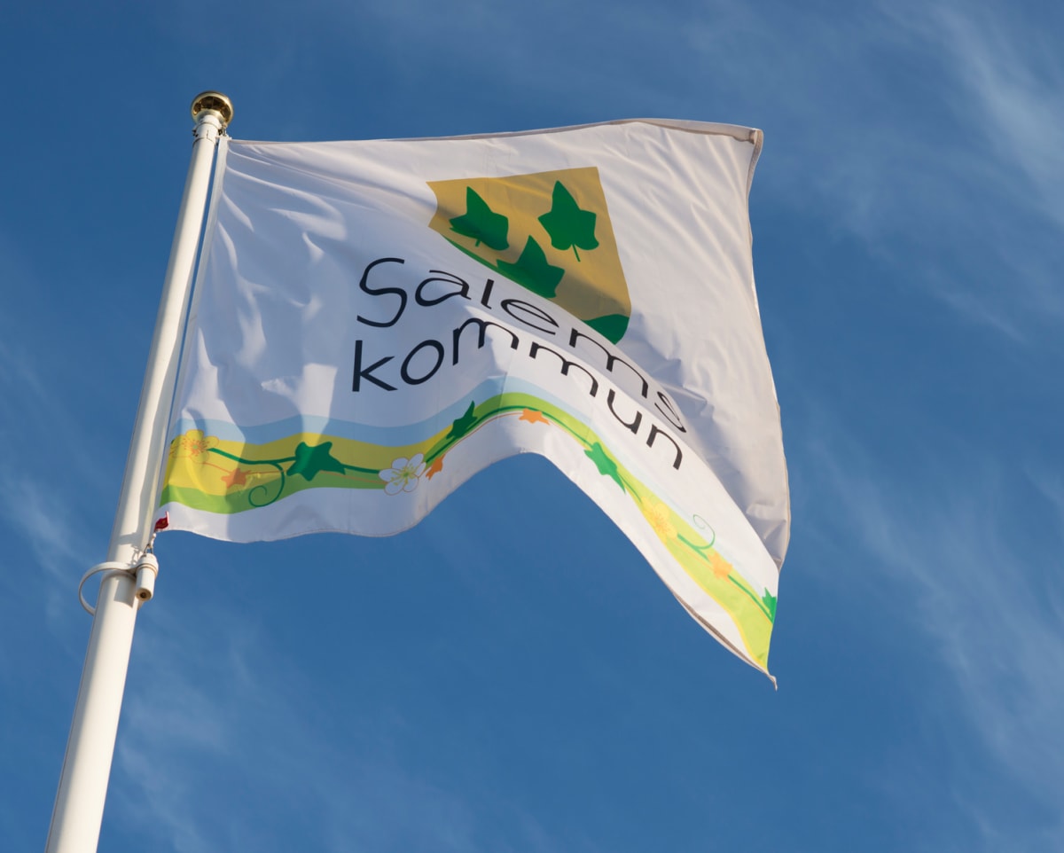 En vit flagga med texten "Salems kommun" och en grön-gul symbol med blad, samt ett horisontellt band med blommor och blad. Flaggan är fäst vid en vit flaggstång och vajar mot en klarblå himmel med tunna, vita moln.