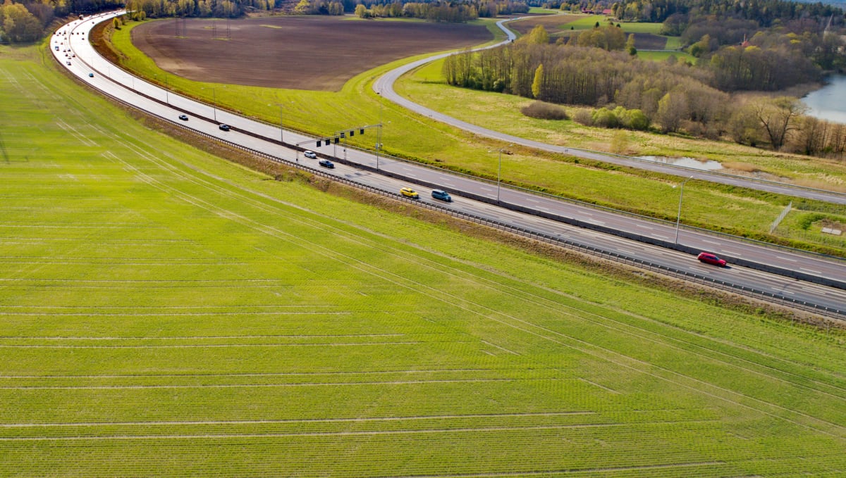Flygfoto över en motorväg med ett flertal bilar som kör i båda riktningarna. Runt vägen finns gröna fält och träd, och i bakgrunden syns ett skogsområde och en sjö. Två motorvägar korsar varandra.