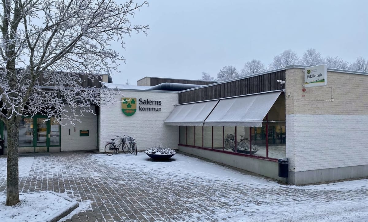 En exteriör vy av en byggnad med snö på marken och träd. Fasaden är vit med en mörkare sektion upptill, och en skylt med texten "Salems kommun" och "Bibliotek" är synlig. Två cyklar står parkerade utanför och snö ligger på marken och byggnadens tak.