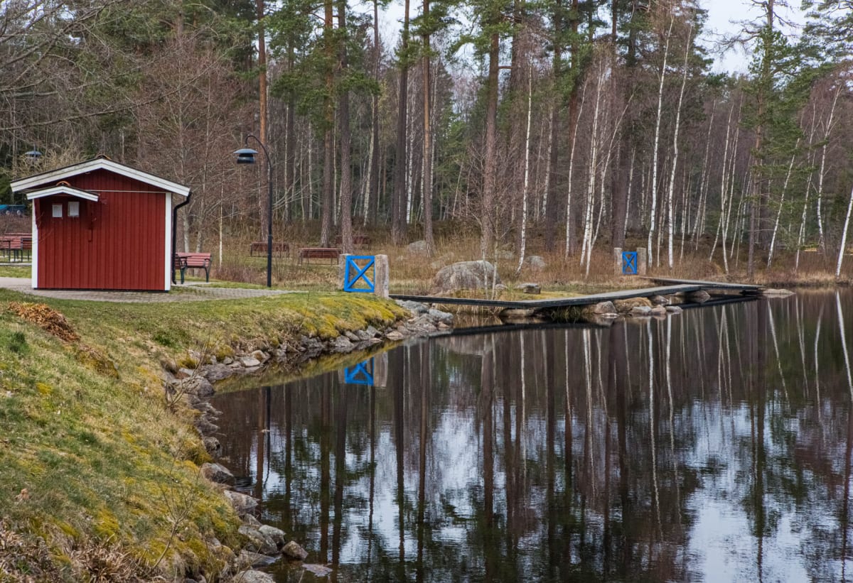 En bild av en röd stuga med vita knutar vid en sjö eller tjärn. Bredvid stugan finns en gräsyta, en asfalterad gång, och en parkbänk. I bakgrunden syns en skog med lövfällande träd och barrträd. En träbrygga sträcker sig ut över vattnet där träden re