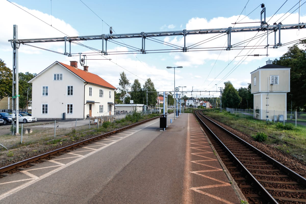 En bild som visar en järnvägsstation med spår, en perrong och en vit byggnad med rött tak. I bakgrunden syns ett tågspår som svänger, samt ett gulmålat torn och bostadshus. Himlen är delvis molnig och lövträd är synliga.