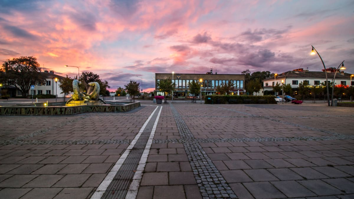 En bild tagen i skymningen med en rosa-lila himmel över en stadsbild. På förgrunden syns ett stort torg belagt med marksten. Till vänster i bilden finns en upplyst fontän och i bakgrunden syns flera byggnader och träd. Gatlyktor är tända och belyser