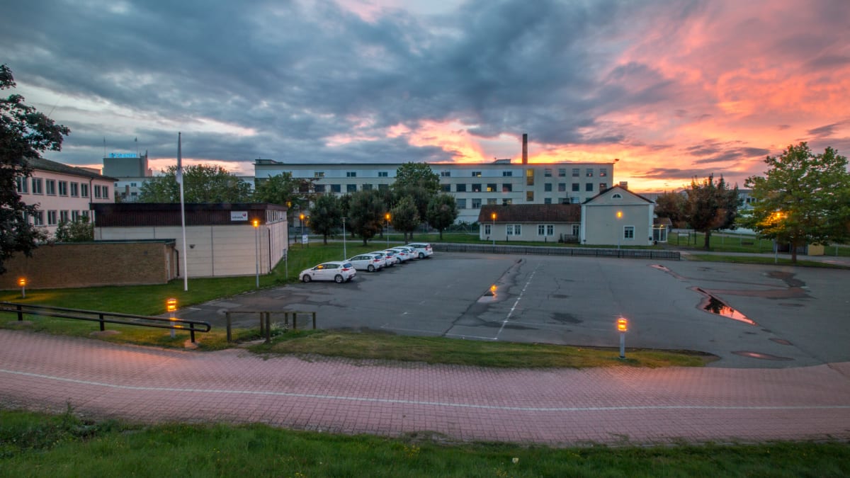 En översiktsbild av en parkeringsplats med flera vita bilar, omgiven av byggnader och träd, under en solnedgång med orangefärgad himmel och moln. Det finns gatlyktor tända och vattenpölar på asfalten.