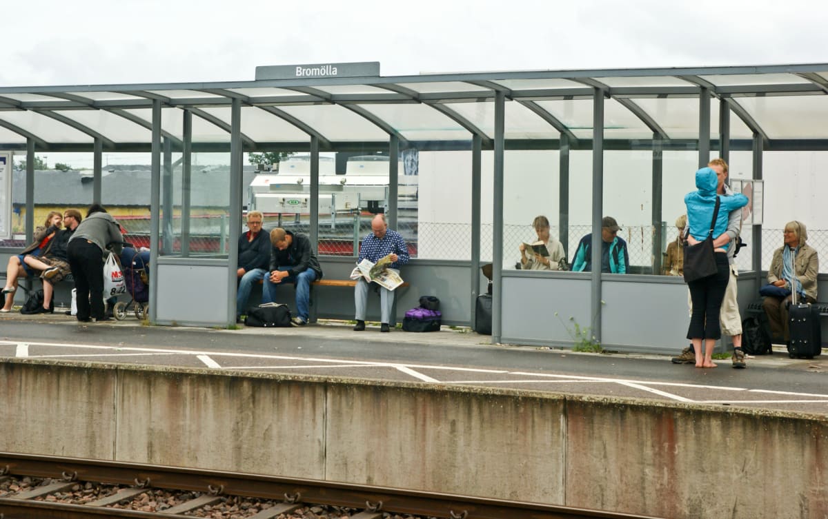 En grupp människor väntar på en perrong vid en tågstation under ett väderskydd. En skylt med texten "Bromölla" är synlig. Flera personer sitter på bänkar medan andra står. En barnvagn står i bakgrunden. Ett tågspår syns i förgrunden.