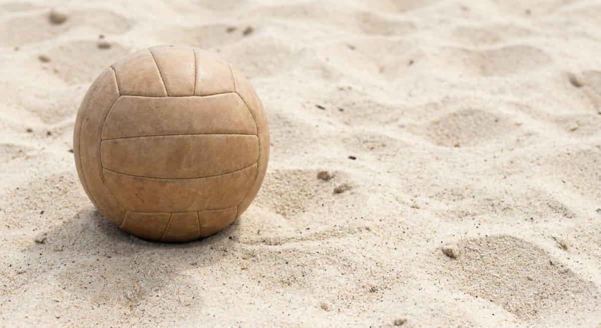 En beige volleyboll ligger till vänster i bilden, delvis nedsjunken i ljus sand med synliga korn och ojämnheter. Större delen av bilden upptas av sand.