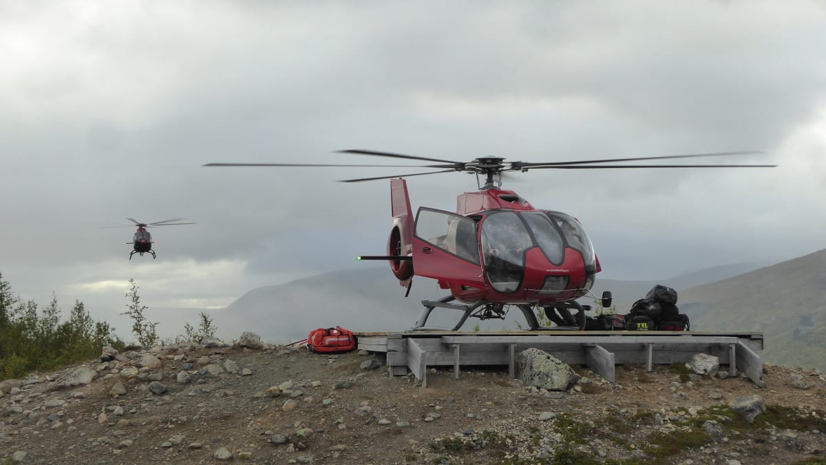 En röd helikopter står parkerad på en träplattform på en stenig kulle. En annan helikopter syns i luften i bakgrunden. Bergen i bakgrunden är delvis dolda av moln eller dimma. Kebnekaise fjällstation