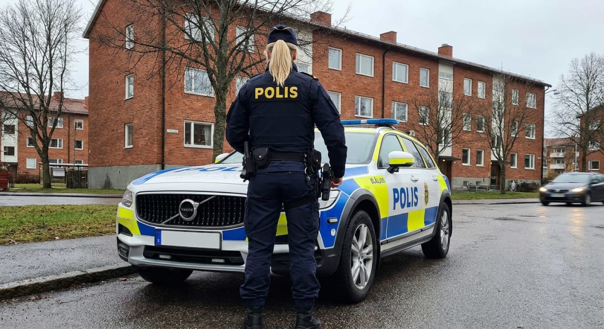 En person i polisuniform står vänd bort från kameran framför en svensk polisbil. Bilen är parkerad på en gata med röda tegelbyggnader och träd med kala grenar i bakgrunden. Vägen är våt och en annan bil skymtar i rörelse.