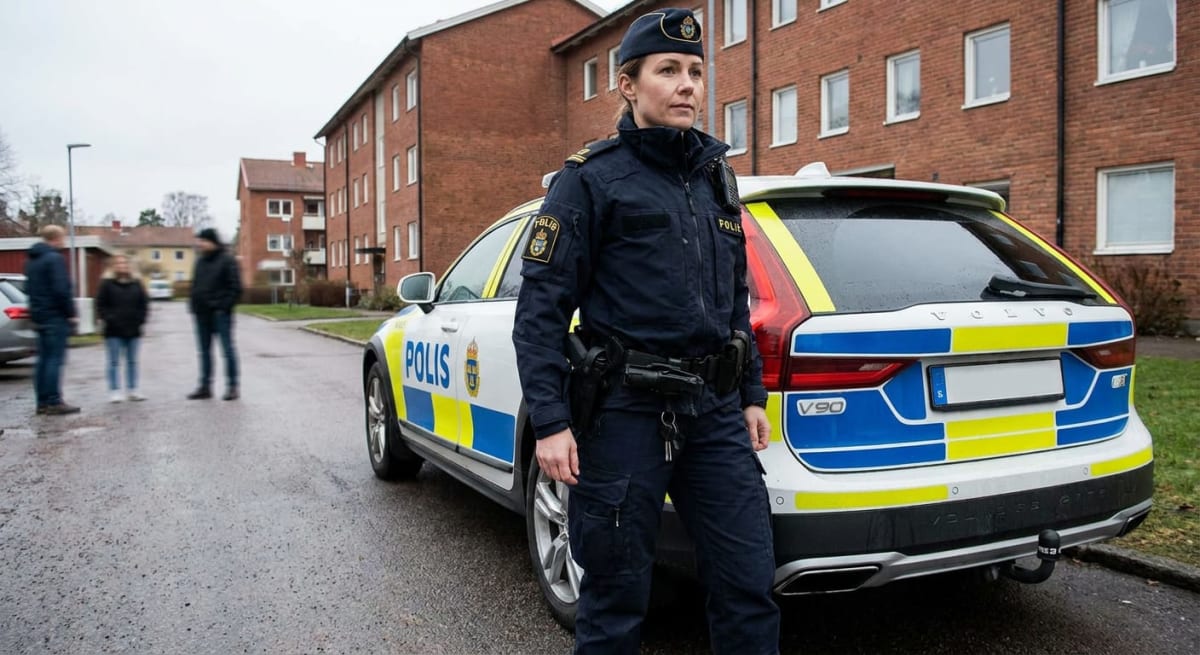 En person i svensk polisuniform står vid en polisbil (Volvo V90) på en asfalterad yta framför flerfamiljshus i tegel. Uniformen inkluderar en keps, en mörkblå jacka och byxor med polismärken samt bälte med utrustning. Ytterligare tre personer syns i