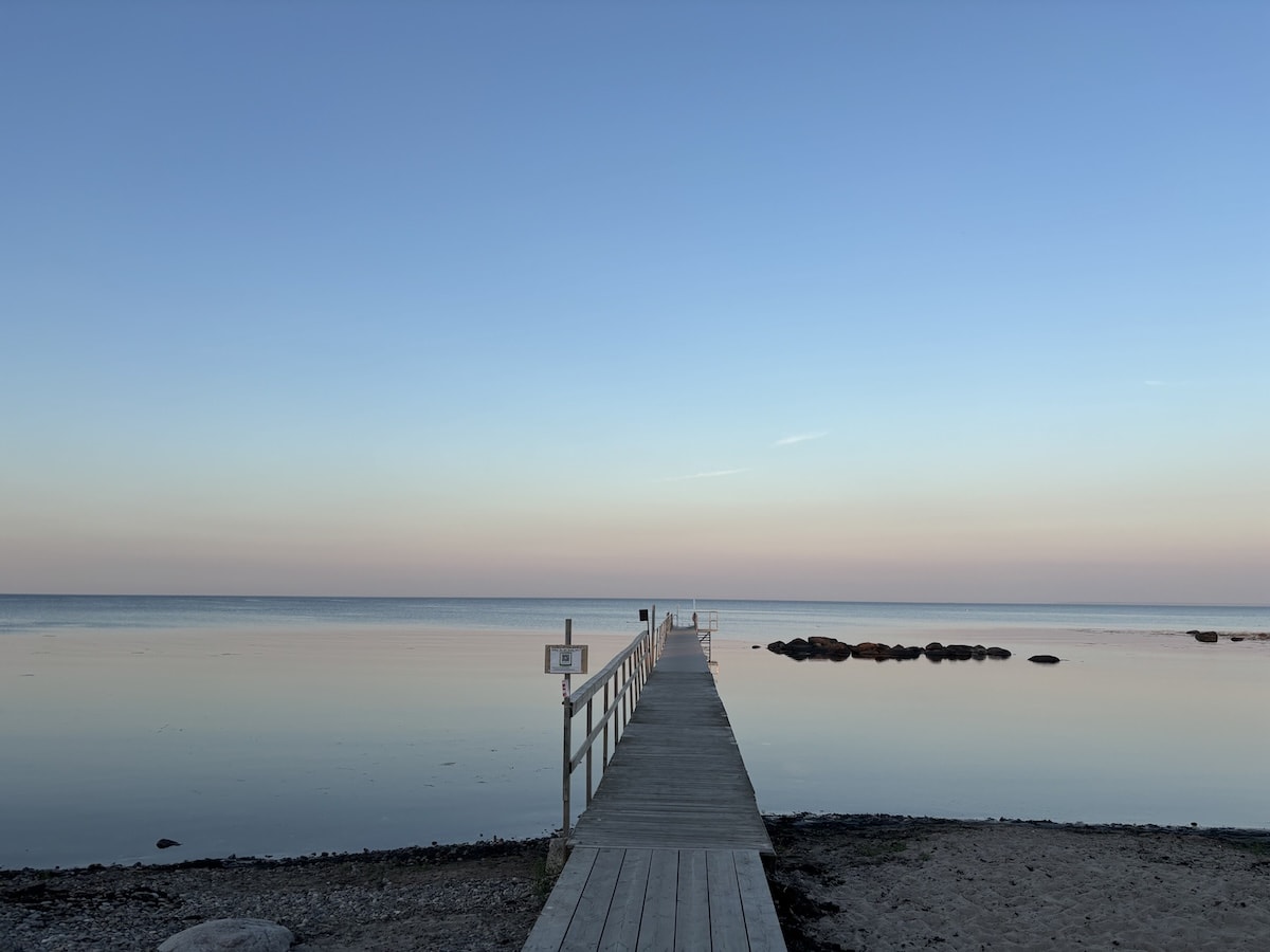 En träbrygga sträcker sig ut över lugnt vatten vid en strandkant med sand och stenar. Horisonten visar en molnfri himmel som övergår från ljust blått till en svag rosa ton nära horisonten. Några stenar syns i vattnet en bit ut från stranden.