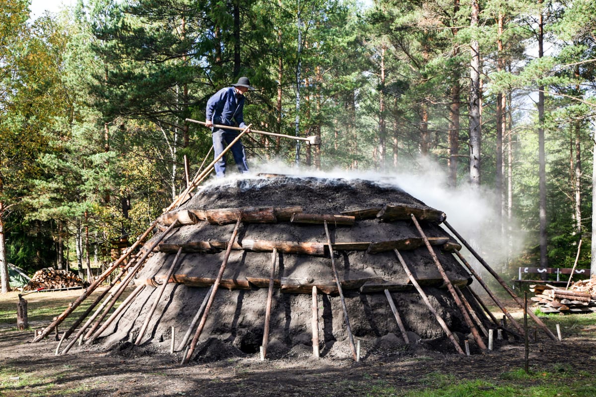 En person i hatt och blå kläder står på toppen av en kolmila som avger rök, omgiven av en grön skog. Personen hanterar ett långt verktyg. I bakgrunden syns travade vedhögar och en bänk.