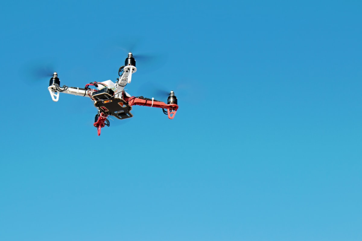 En fyrpropellerdrönare med vita och röda armar ses flyga mot en klarblå himmel. Propellrarna snurrar och drönarens underrede med elektronik och kablar är synligt.