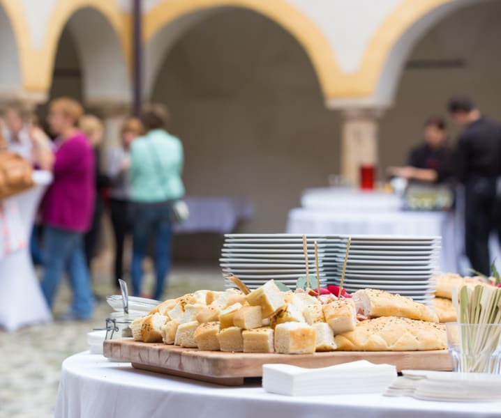 Bilden visar ett evenemang utomhus där mat och dryck serveras. I förgrunden står ett vitt dukat bord med smörgåsbitar och bröd upplagt på en träskärbräda. Bredvid brödet finns staplar med vita tallrikar och små träspett. I bakgrunden syns suddiga män