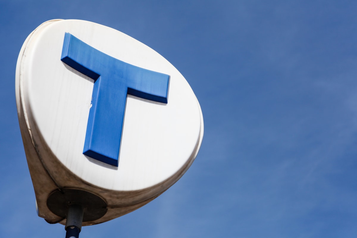 En vit, rund skylt med ett blått versalt "T" i mitten, monterad på en stolpe, syns mot en klarblå himmel. Skylten är smutsig längs kanterna och det blåa "T:et" har vissa ojämnheter. Solen lyser från vänster och kastar skugga på skylten.