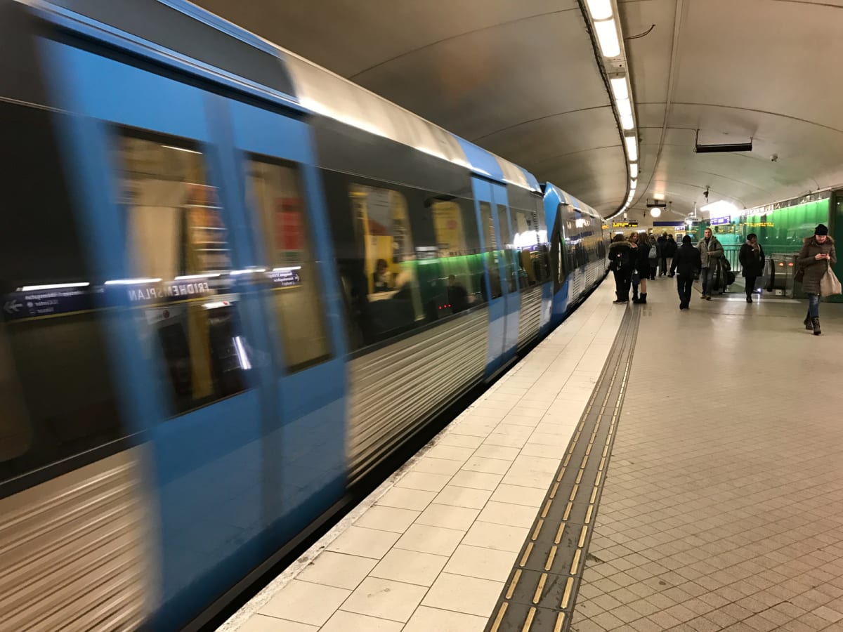 En blå och silverfärgad tunnelbanevagn rör sig längs en perrong i en underjordisk station. Flera personer står och går på perrongen, som är upplyst av lysrör i taket. På tågets sida syns en spegelvänd skylt med texten "FRIDHEMSPLAN".