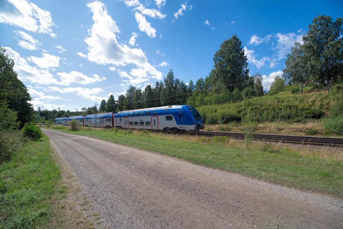 En blå och vit flerdelad pendeltågsliknande tågvagn från mälartåg kör på en järnväg bredvid en grusväg. Omgivningen består av grön vegetation och träd, samt en ljusblå himmel med vita moln.