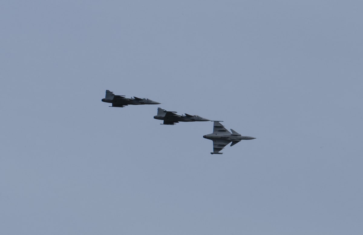 Tre gråa militära stridsflygplan, av typen JAS 39 Gripen, flyger i formation mot en nästan molnfri ljusblå himmel. Ett flygplan är närmast betraktaren och två mindre synliga längre bort, placerade diagonalt i bilden.