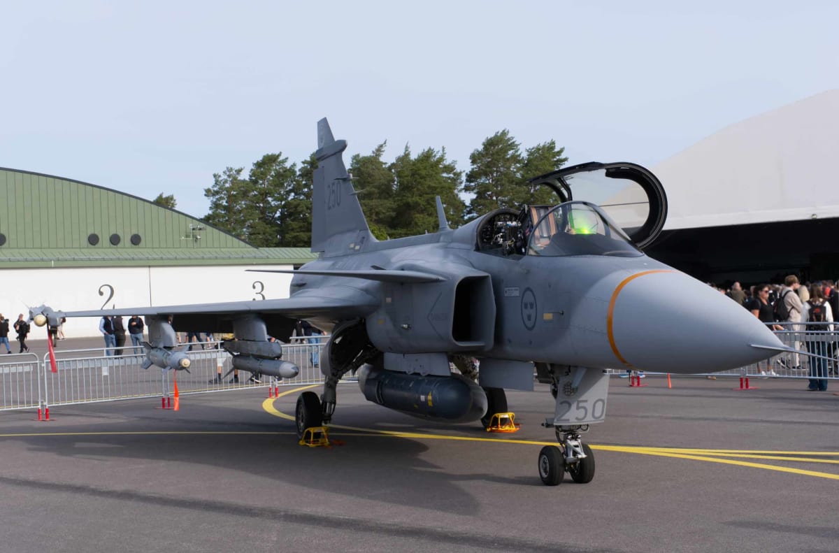 En grå stridsflygplan av typen Saab Gripen står på asfalt med sitt landningsställ. Flygplanets cockpit är öppen och flertalet externa vapen och bränsletankar är monterade under vingar och flygkropp. En folkmassa syns i bakgrunden bakom ett stängsel,