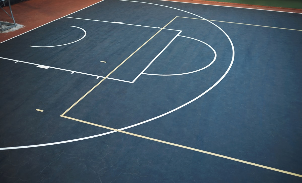 En yta med mörkblått underlag som ser ut att vara en del av en basketplan. Vita och gula linjer markerar olika spelområden. I bildens övre vänstra hörn syns en rödbrun yta och en del av en metallkonstruktion.