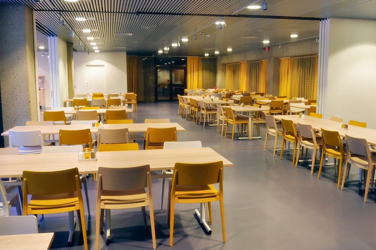 En ljus inomhusmiljö med många tomma bord och stolar i rader, en skolmatsal eller skol-cafeteria. Väggarna är ljusa och taket har infälld belysning. Gula gardiner syns längs sidoväggarna.