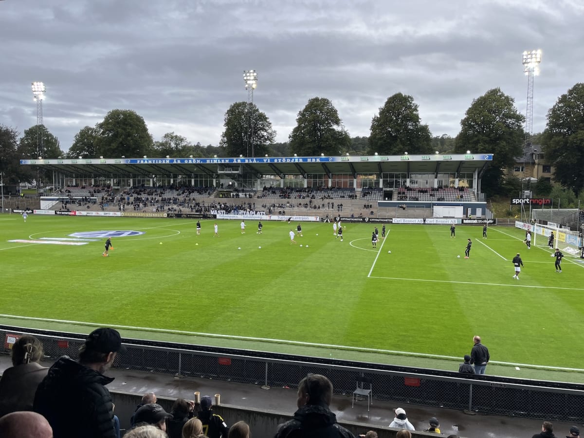 En översiktsbild av en fotbollsarena med en grön fotbollsplan där spelare tränar. En stor läktare med publik och reklamskyltar syns i bakgrunden under en mulen himmel. Flera strålkastartorn står upplysta.
