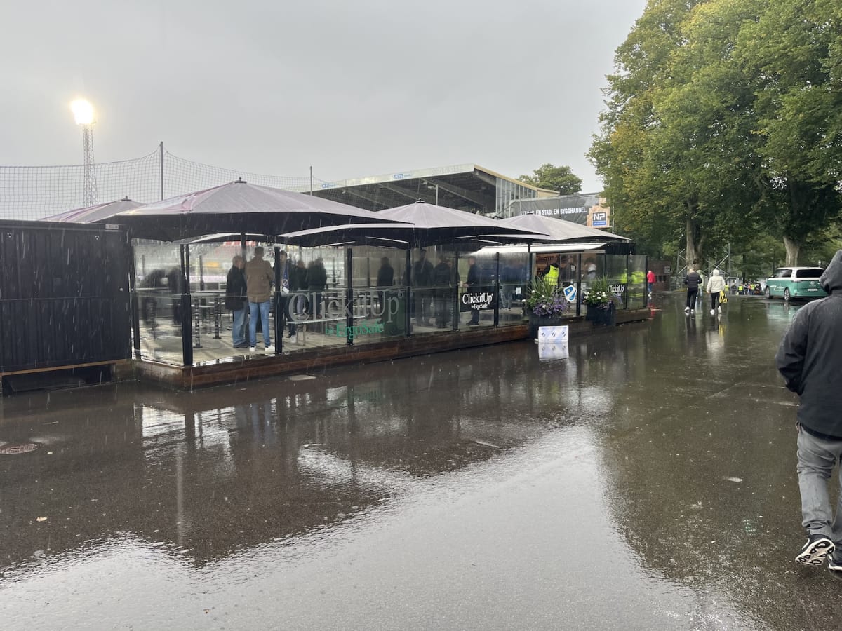 En utomhusbild tagen under en regnig dag visar en uteservering med svarta parasoller och glasväggar. Människor syns stå under parasollerna och på den blöta asfalten. I bakgrunden syns ett fotbollsmål, en strålkastare och grönskande träd. Marken är tä