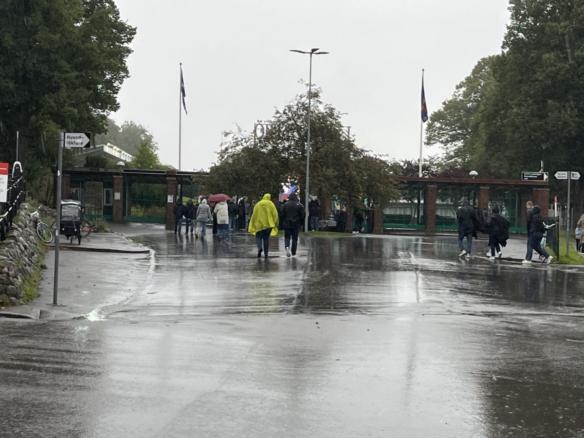 En bild tagen utomhus under dagtid i regnigt väder visar en asfalterad yta med vattenpölar, framför en ingång med tegelpelare och två flaggstänger med flaggor. Flera personer, varav en bär en gul regnjacka, går eller står i bildens mitt och bakgrund.