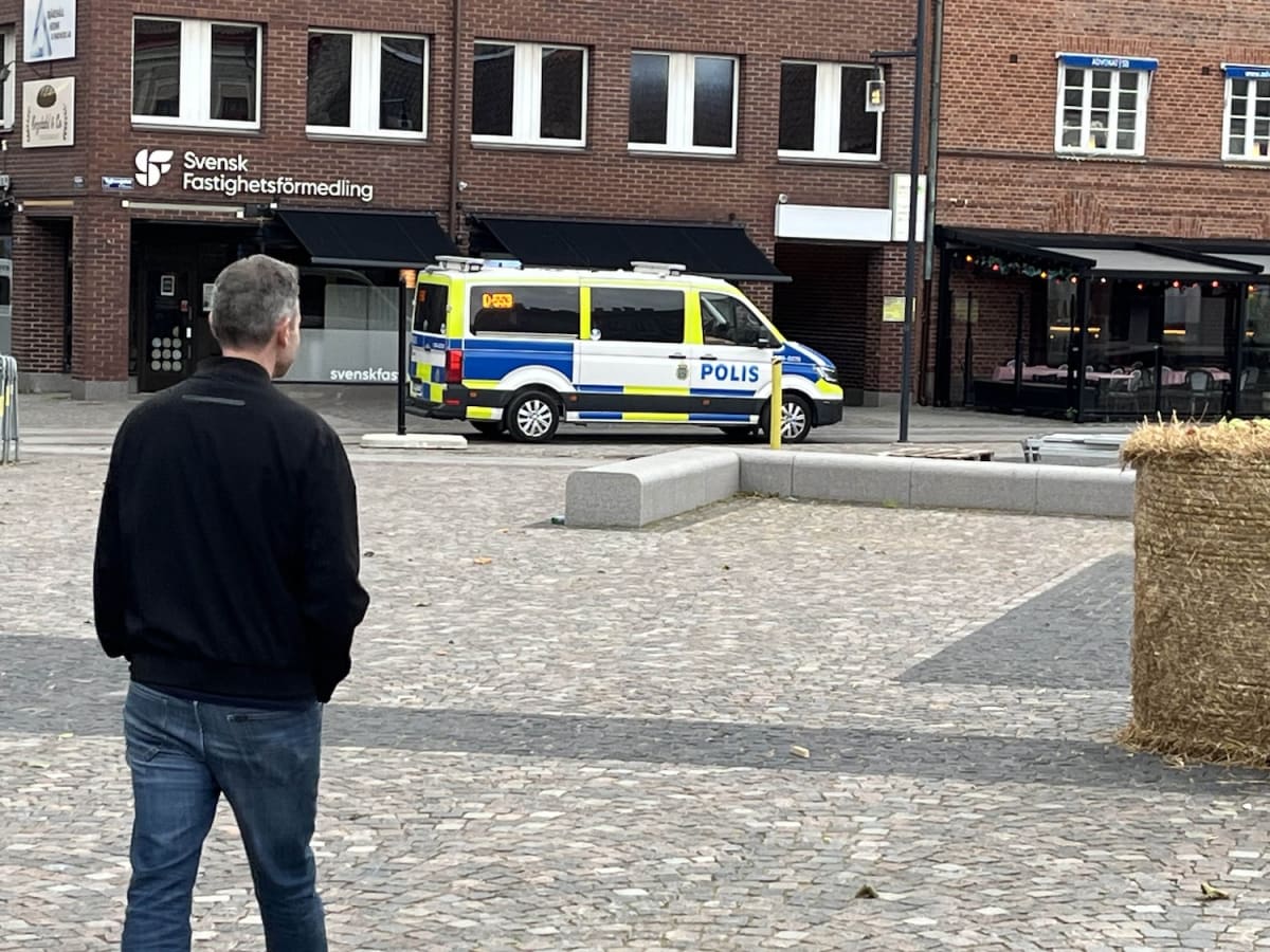En person sedd bakifrån står på ett torg med kullersten. En svensk polisbil är parkerad på gatan i bakgrunden, framför en byggnad med skylten "Svensk Fastighetsförmedling". Till höger syns en restaurang med uteservering och en halmbal.