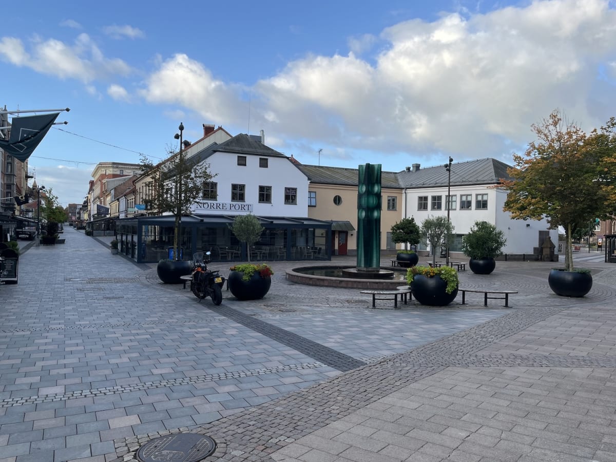 En bild över ett tomt torg med en fontän och en grön skulptur i mitten. Runt torget syns byggnader, träd med höstfärgade löv och restauranger med uteserveringar. En motorcykel står parkerad nära en av byggnaderna. Himmelen är blå med vita moln.
