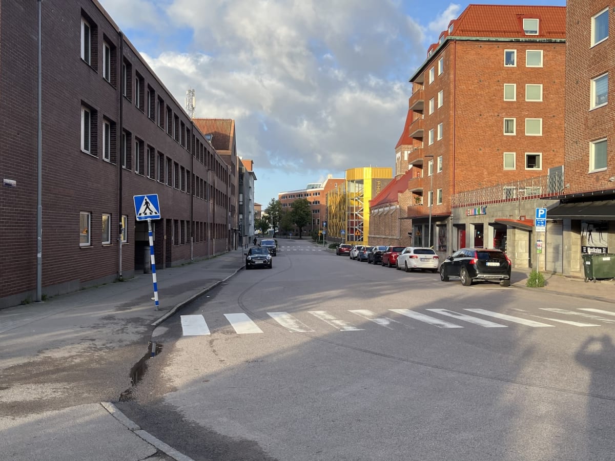 En gata med parkerade bilar längs ena sidan och en fotgängare skylt synlig på den andra. Byggnader i tegel och en gul byggnad med trappor syns. Ett övergångsställe är målat på asfalten under en delvis molnig himmel.