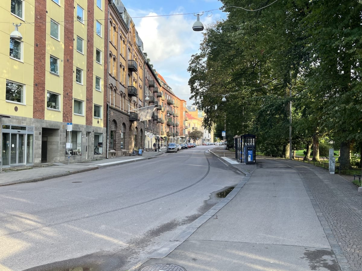 En gata kantad av flerbostadshus på ena sidan och en park med träd på den andra. På gatans vänstra sida syns parkerade bilar och på höger sida finns en busskur och en gångväg. Himmelen är ljus med moln och det är dagsljus.