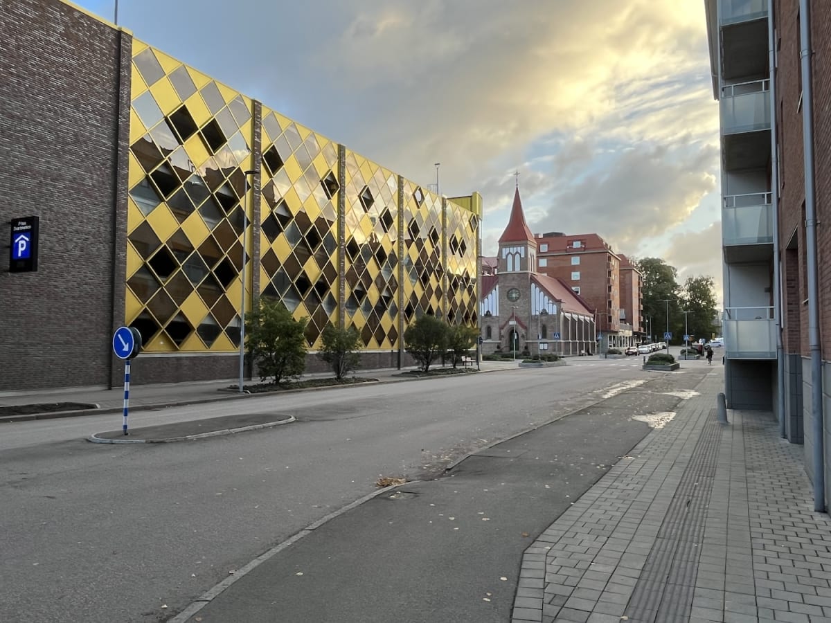 En gata med ett stort parkeringshus med guldfärgade rutor på vänster sida och bostadshus i tegel på höger sida. Längre bort syns en kyrka med rött tak. Himlen är delvis molnig med inslag av blå himmel och ljusa moln. Asfalt, trottoarer och viss veget