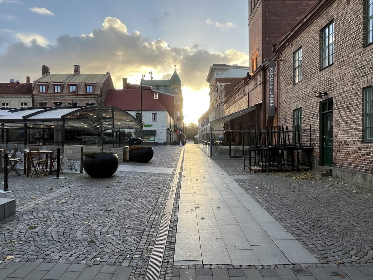 En gata i en stad kantad av tegelbyggnader med fönster och uteserveringar. Solen lyser lågt på himlen och skapar långa skuggor och ett gyllene ljus över molnen. Gatubeläggningen består av kullersten och stenplattor, och det ligger några enstaka löv p