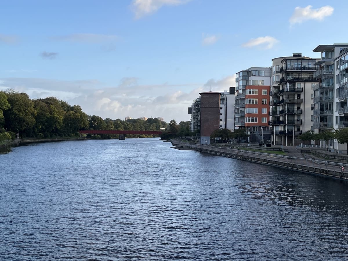 En bild som visar en flod eller å med bebyggelse på höger sida och trädklädda stränder på vänster sida. I bakgrunden syns en röd bro över vattnet. Himlen är delvis molnig med blåa partier och byggnaderna är moderna flerfamiljshus med balkonger.
