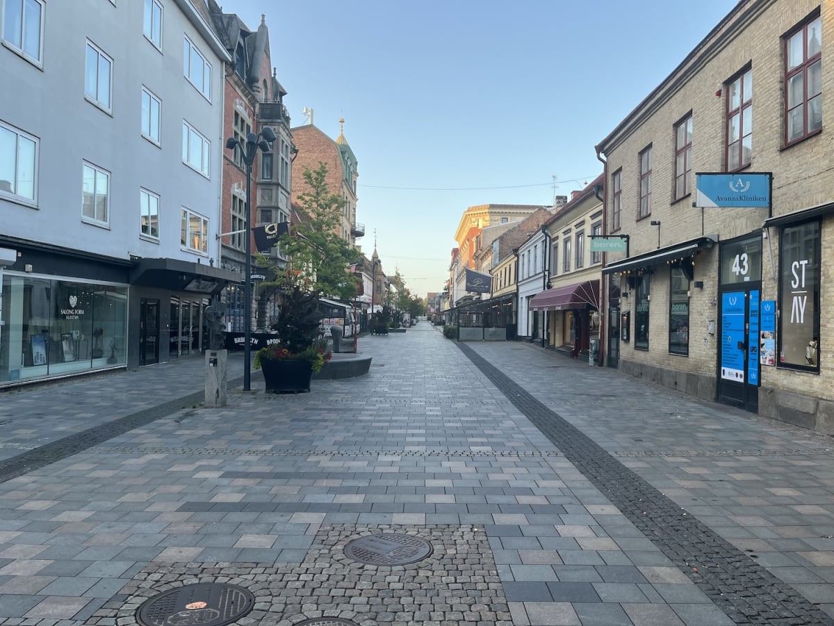 En tom gågata kantad av flervåningshus med butiker och restauranger på båda sidor. Gatsten täcker marken och belysningsstolpar syns längs gatan. Himlen är klar och ljus. Träd och planteringar med grönska och blommor syns på flera platser.