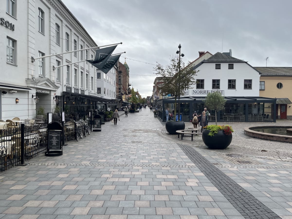 En gata med stenbeläggning, flankerad av byggnader med restauranger och uteserveringar. Flera personer promenerar längs gatan och sitter vid bord. Molnig himmel syns ovanför.