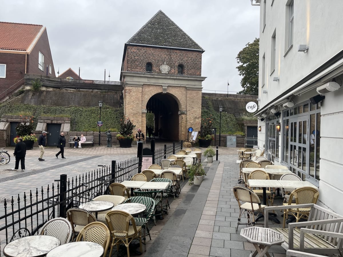 En gata med gatsten framför en tegelbåge med torn som leder under en befästningsvall. På höger sida finns en restaurang med uteservering och många tomma bord och stolar. Ett fåtal personer syns på gatan och en bänk. Himlen är grå och molnig.